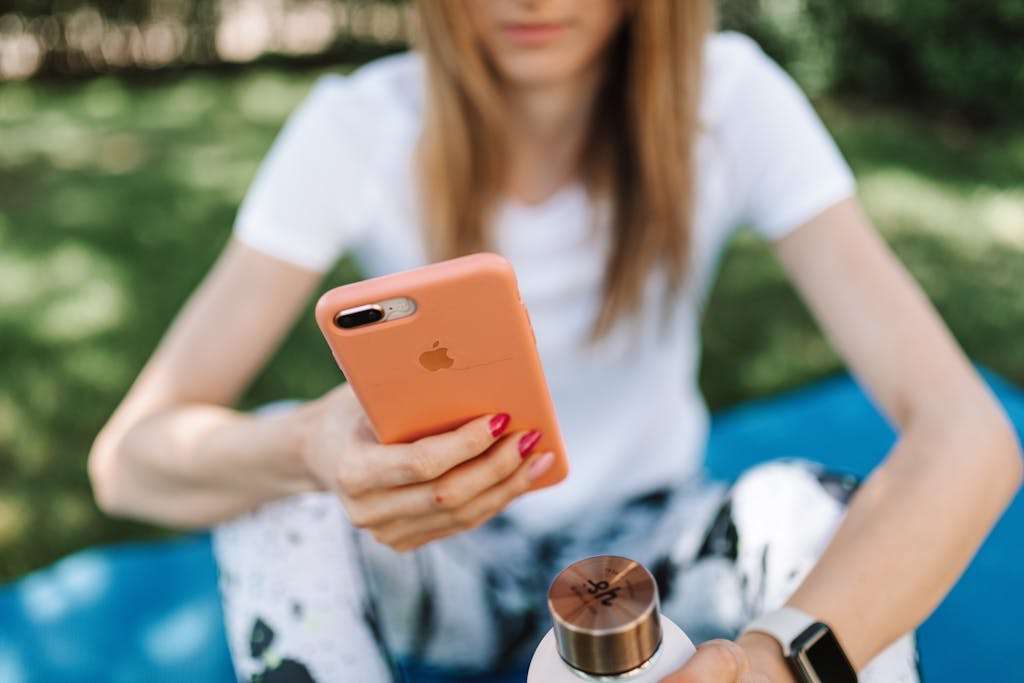 A young woman seated on a mat using her smartphone outdoors in a sunny park.